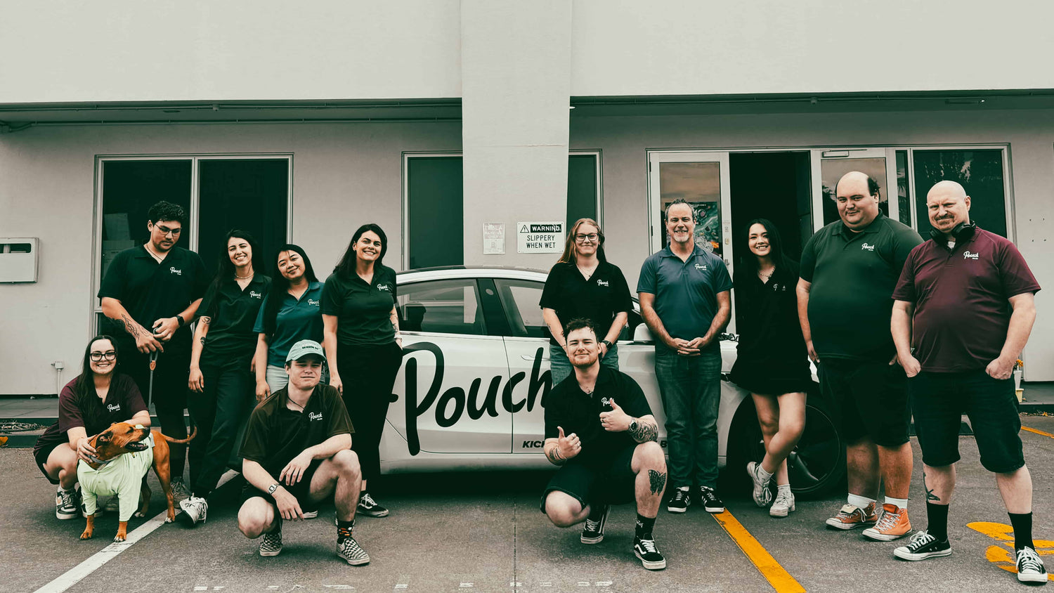 Group of people posing in front of a building with a car branded 'Pouch'.